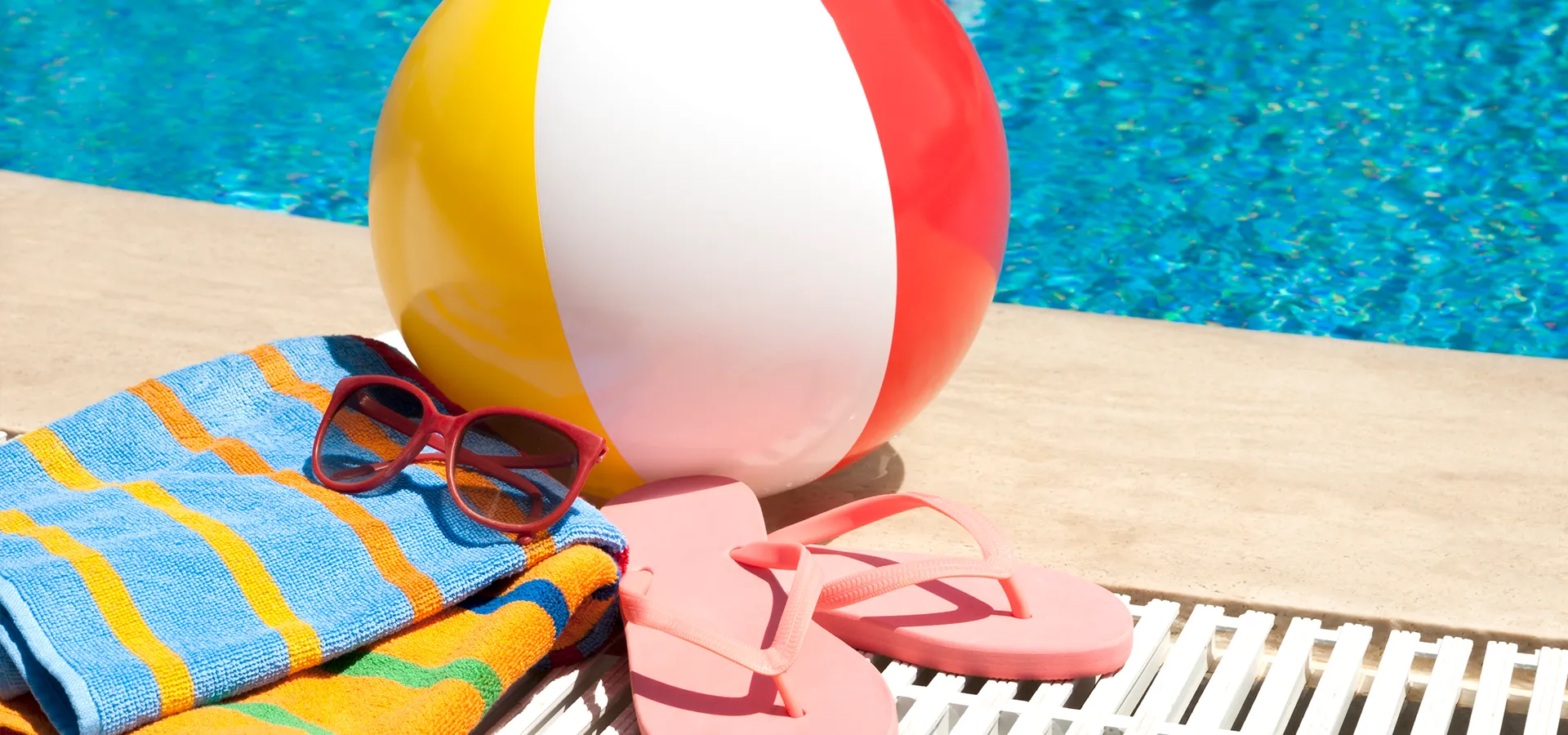 ein Badetuch, Flip Flopsund ein Wasserball liegen am Beckenrand